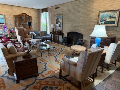 A living room of antique and upholstered furniture over a patterned rug, with framed paintings and a china cabinet against the far wall